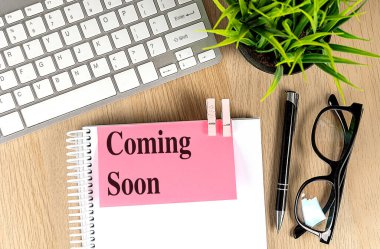 COMING SOON text pink sticky on a notebook with keyboard, pen and glasses .  clipart