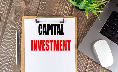 Diz üstü bilgisayarı, faresi ve kalemi olan bir pano kağıdının üzerindeki CAPITAL INVESTMENT metni . 