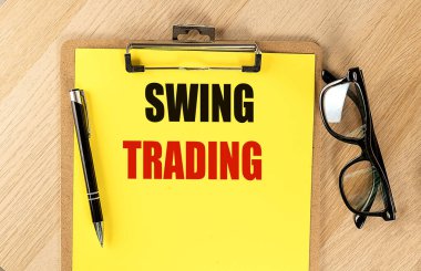 SWING TRADING text on a yellow paper on clipboard with pen and glasses.  clipart