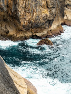 Dalgalar kayalık kayalıklara çarpıyor, beyaz köpük ve engebeli kıyı manzarası yaratıyor. . 