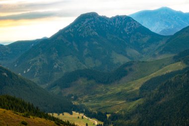 Dönek yolu ve sık ormanları olan dağ vadisi alacakaranlıkta bulutlu bir gökyüzünün altında yükselen tepelerle çevrilidir. . 