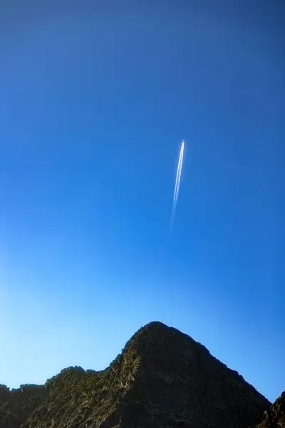 stock image  Airplane flying high in clear blue sky above a rugged mountain peak, leaving a white contrail behind . 