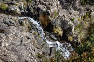 Çimen ve bitki örtüsüyle çevrili engebeli kayaların üzerinden akan küçük şelale. . 