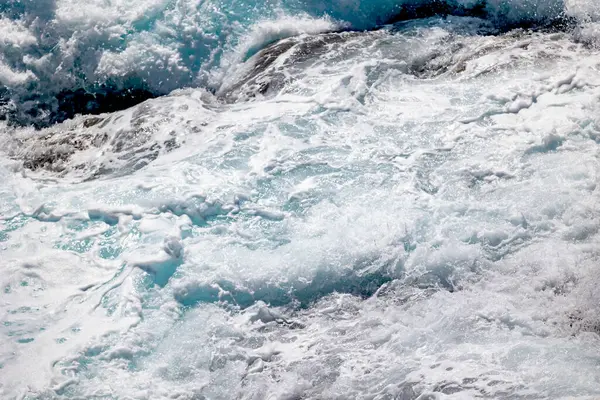 Güneş ışığında kıyı kayalarının üzerine düşen köpüklü turkuaz deniz dalgalarının yakın çekimi . 