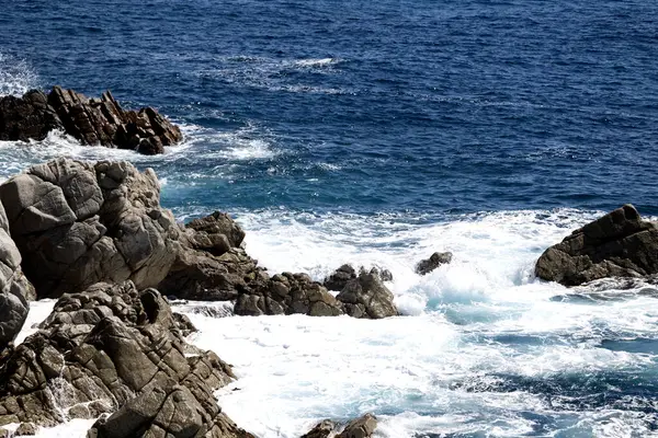 Parlak güneş ışığı altında parlak mavi kıyı şeridi boyunca sivri kayalıklara köpüklü dalgalar çarpıyor.. 
