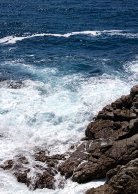 Güneşli sahil gününde köpüklü turkuaz dalgalar engebeli kayalara çarpar. 