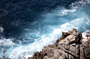 Güneş ışığı, kıyı kayalarına çarpan turkuaz bir deniz dalgasında parıldıyor. . 