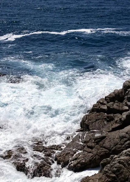 Güneşli sahil gününde köpüklü turkuaz dalgalar engebeli kayalara çarpar. 