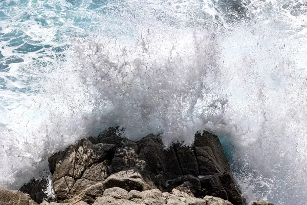 Büyük bir okyanus dalgası kıyı kayalarına çarpıyor ve dramatik bir şekilde beyaz köpük sıçrıyor. . 