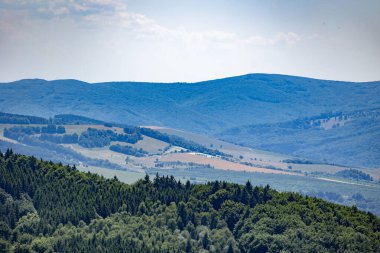 Yeşil ormanları ve ekili tarlaları olan yuvarlanan arazi uzak mavi dağlar boyunca uzanıyor. . 