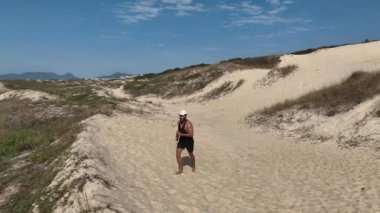 Sahildeki kum tepelerinde yürüyen ve koşan bir adam. Adamı takip eden İHA 'lı hava görüntüsü. Yukarıdan görüntü. Sağlık kavramı.