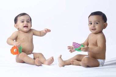 İkiz bebekler, parlak oyuncaklarla yan yana oynuyorlar. Stüdyo zemininde otururken gülümsüyorlar. Arkadaşlık, çocukluk bağı ve aile teması için mükemmel..