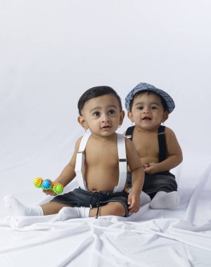 İkiz bebekler birbirine yakın oturuyor, biri renkli oyuncaklar taşıyor, ikisi de pantolon askısı takıyor, aile bağlarını, çocukluk oyunlarını ve arkadaşlığı simgeleyen sevimli bir portre..