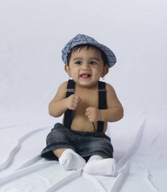 Bebek çocuk mavi kareli şapka ve pantolon askısı takıyor, sade stüdyo arka planında otururken mutlu bir şekilde gülümsüyor, bebek modası ve çocukluk için mükemmel.