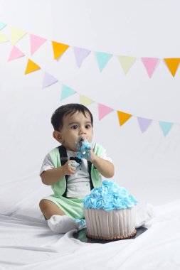 Yeşil kıyafetli şirin bir bebek pasta ezme kutlamasını seviyor. Arka planda parti havası olan büyük mavi bir kekin önünde oturuyor. Çocukluk teması için mükemmel..