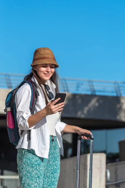 Tourist woman smiling while using her mobile phone standing outdoors with suitcase. Travel, tourism and technology concept.