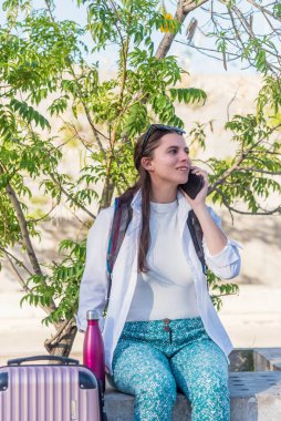 Dışarıda bavuluyla oturan bir turist kadın telefonda konuşuyor. Teknoloji ve seyahat kavramı.