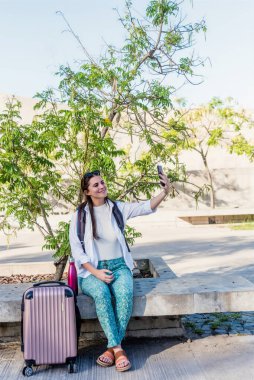Genç turist kadın dışarıda bavuluyla otururken cep telefonuyla selfie çekiyor. Teknoloji ve seyahat kavramı.