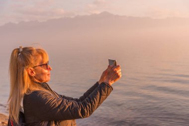 Gün batımında doğal göl manzarasını fotoğraflayan cep telefonlu olgun bir kadın. Teknoloji ve seyahat kavramı.