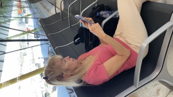 Reife Blonde Frau Sitzt Auf Flughafenbank Und Tippt Auf Smartphone — Stockvideo