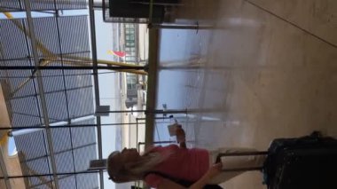 Olgun bir kadın havaalanı koridorunda yürüyor. Elinde bir şişe su olan yuvarlanan bir bavul ile kapıyı arıyor..