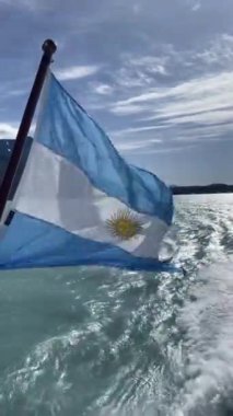 Suda sarı güneşli Arjantin mavi ve beyaz bayrağı dalgalanıyor. Bayrak bir direğe bağlı ve su tarafından taşınıyor. Huzurlu ve huzurlu bir sahne.