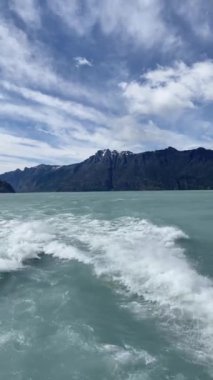 Bir teknenin arkasından görülen Lago Argentino 'nun suyu mavidir ve gökyüzü bulutludur. Arkadaki dağlar karla kaplı..