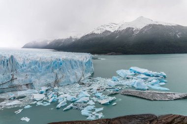 Perito Moreno 'nun büyük bir buz kütlesi buzdağları ve karlarla çevrilidir. Gökyüzü bulutlu ve arka plandaki dağlar karla kaplı. Sahne huzurlu ve huzurlu, buzlu.
