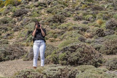 Bir kadın kayalık bir yamacın fotoğrafını çekiyor. Tepenin yamacı çalılarla kaplı ve kayalık ve çorak bir görünüşü var. Kadın siyah gömlek ve beyaz pantolon giyiyor.