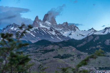 A mountain range with snow on the peaks and a cloudy sky. The mountains are in the background and the sky is blue