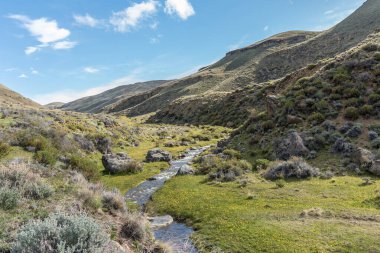Bir nehir, üzerinde açık mavi gökyüzü olan çimenli bir vadiden geçiyor. Sahne huzurlu ve sakin, su yavaşça akıyor ve çimenler doğal bir zemin sağlıyor.