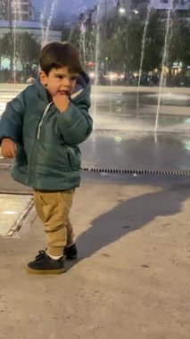 Bir çocuk çeşmenin önünde yürüyor ve zıplıyor ve rutubetin ortasında gülüyor. Yeşil ceket ve kahverengi pantolon giyiyor..