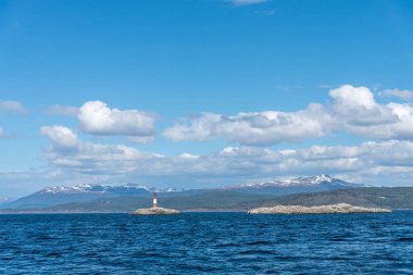 Les Eclaireurs 'un kayalık bir adadaki deniz feneri, Beagle Kanalı' nın mavi suları ve Arjantin 'in Ushuaia yakınlarındaki açık mavi gökyüzünün altındaki uzak karlı dağlarla çevrili.. 