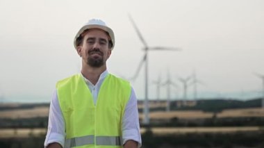 Açık hava rüzgar türbini parkında kask ve yelek giyen genç bir mühendisin portresi. Mühendislik ve güvenlik iş başında