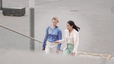 İki iş arkadaşı merdivenlerden çıkıp ofise doğru yürüyorlar, sohbet ediyorlar, sarışın ve Asyalı profesyonel kadınlar.