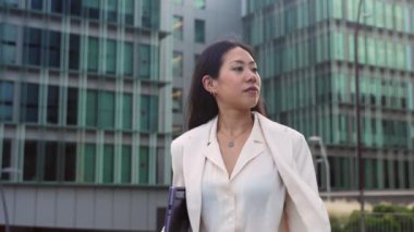 Tablet bilgisayarlı başarılı genç Asyalı kadın iş binalarının yanında yürüyor ve neşe içinde gülümsüyor. Zarif yönetici kız. 