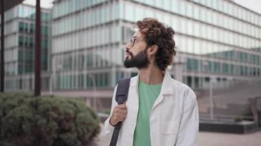 Sakallı, gözlüklü, şehirde yürüyen ve kameraya gülümseyen kendinden emin genç profesyonel adam. 