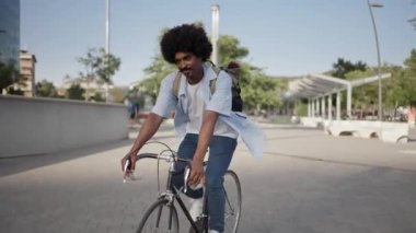 Young African American hipster man with bag riding bicycle on city street going to work 