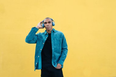 Mavi ceketli Afro-Amerikan adam kulaklıkla müzik dinlerken, sarı arka planda el ele dururken. 