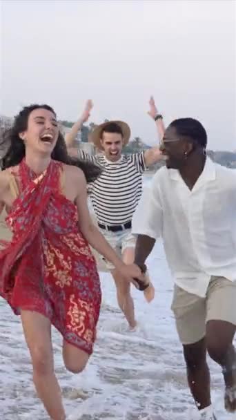 幸せな友人裸足で一緒にビーチに沿って実行している水の波の中で日没スプラッシュ 旅行夏休み 垂直ビデオ — ストック動画