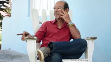 Yaşlı Kübalı adam puro içiyor, verandada oturuyor ve başka tarafa bakıyor.
