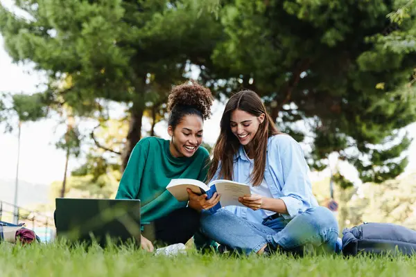 Women students sitting on public park grass learning together with book and laptop, studying for University exams