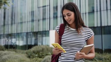 Üniversite kampüsünde sarı cep telefonu kullanan mutlu İspanyol öğrenci kız. Gülümseyen genç kadın çevrimiçi uygulamayla eğleniyor 