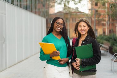 Üniversiteli iki kız üniversite kampüsünde kameraya bakıp gülümsüyor. Asya ve Afrika Amerikan portresi
