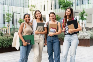 Dört genç kız öğrenci okul önünde kitap tutarak kaynaşıyorlar.