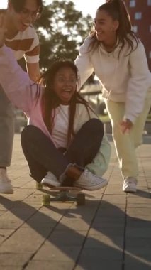 Neşeli Afrikalı Amerikalı kız kaykaya biniyor Asyalı genç bir arkadaş tarafından itiliyor, şehirde kaykay yaparken eğleniyor - arkadaşlık oyunları