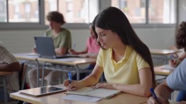Üniversite öğrencisi kız bir sınıfta notlar yazıyor. Lisede çok ırklı gençler okuyor. 