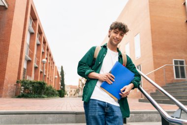 Gülümseyen erkek üniversite öğrencisi kampüs binasında durmuş kameraya bakıp konferansa hazırlanıyor.