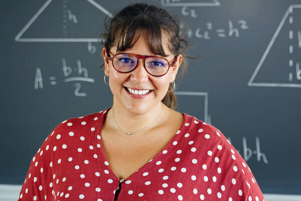 Young female math and geometry teacher standing in a primary school classroom, smiling at the camera