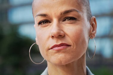 Tıraşlı saçlı şık bir kadın dalgın ve kendinden emin bir ifadeyle kameraya bakar. İnsani duygu ve duygular